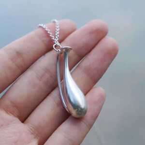 May include: A silver-toned pendant shaped like a slender, elongated vase or teardrop vessel, suspended from a delicate silver-toned chain. The pendant features a smooth, polished metallic surface with a curved, open-frame design.