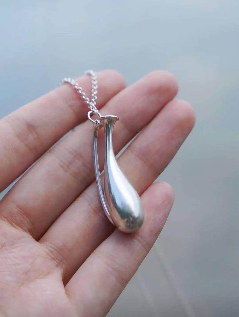 May include: A silver-toned pendant shaped like a slender, elongated vase or teardrop vessel, suspended from a delicate silver-toned chain. The pendant features a smooth, polished metallic surface with a curved, open-frame design.