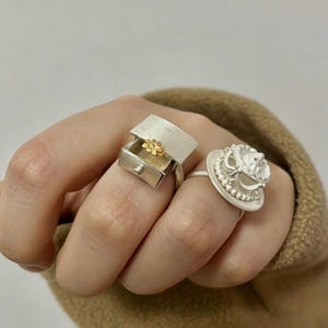May include: Two silver rings. One ring features a miniature silver drawer unit with a small gold-colored flower inside the open drawer. The second ring is a silver circular base topped with a white, ornate, multi-tiered structure with beaded edges.