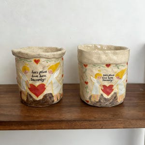 Puede incluir: Dos macetas de cer&aacute;mica beige con un dise&ntilde;o pintado a mano. Cada maceta presenta un coraz&oacute;n rojo, &aacute;ngeles y el texto "Let's plant love here. Secretly!" Las macetas est&aacute;n sobre una estanter&iacute;a de madera.