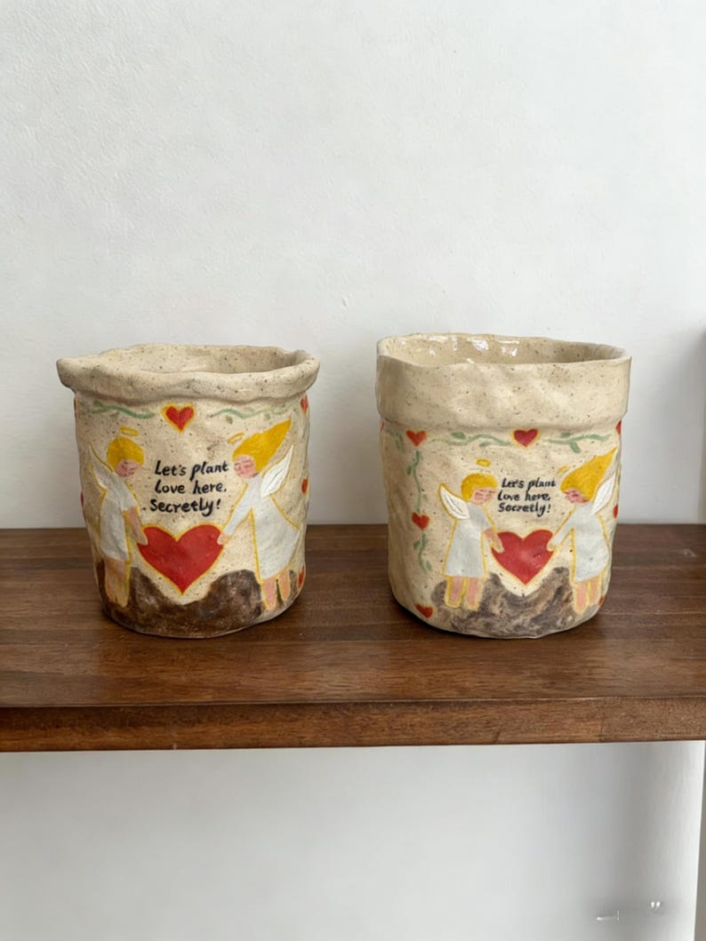 Puede incluir: Dos macetas de cer&aacute;mica beige con un dise&ntilde;o pintado a mano. Cada maceta presenta un coraz&oacute;n rojo, &aacute;ngeles y el texto "Let's plant love here. Secretly!" Las macetas est&aacute;n sobre una estanter&iacute;a de madera.
