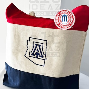 May include: A canvas tote bag with a red top, cream-colored center, and navy blue bottom. The cream section features a navy blue outline of the state of Arizona with the letter "A" inside. The bag has a rope handle and an "Officially Licensed Collegiate Products" logo.