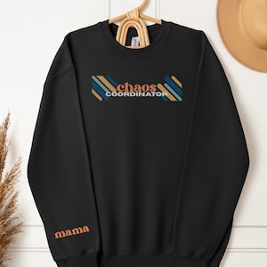 May include: Black sweatshirt with the words "chaos coordinator" in orange and blue text. The word "mama" is embroidered on the left sleeve. The sweatshirt is hanging on a wooden hanger.