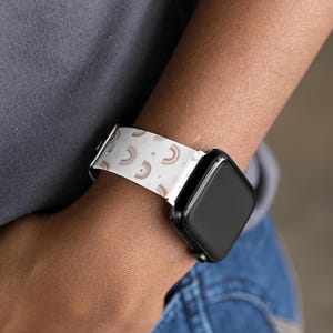 May include: White watch band with a repeating pattern of small rainbows in shades of brown and orange. The watch band is attached to a black smartwatch. The watch band is on a wrist.