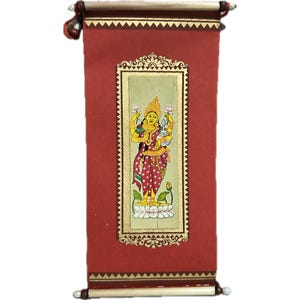 May include: A red scroll painting featuring a detailed illustration of a deity with multiple arms, adorned in colorful garments and jewelry. The artwork is framed with gold accents and set against a red background, with wooden dowels at the top and bottom.