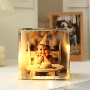 Könnte beinhalten: Ein klarer Acryl-Fotorahmen mit Innenbeleuchtung. Der Rahmen zeigt ein Foto eines Kindes mit Partyhut, das an einem Tisch mit einem kleinen Kuchen sitzt. Ein Holzfotorahmen ist im Hintergrund zu sehen.
