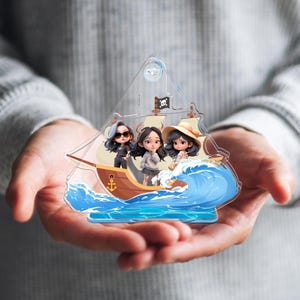 May include: A clear acrylic ornament featuring three cartoon figures on a ship sailing through blue waves. The ship has a pirate flag and a brown hull. The figures are wearing sunglasses, a hat, and casual clothing.