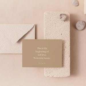 May include: A beige card with the text "This is the beginning of self love. Welcome home." is displayed on a textured, light-colored surface. An envelope and decorative stones are also present, creating a minimalist aesthetic.