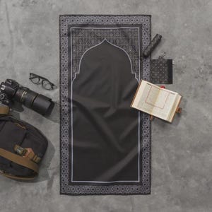 May include: A black prayer mat with a white decorative border and a central arch design. The mat is surrounded by a camera, glasses, a bag, a book, and other accessories. The items are arranged on a gray surface.