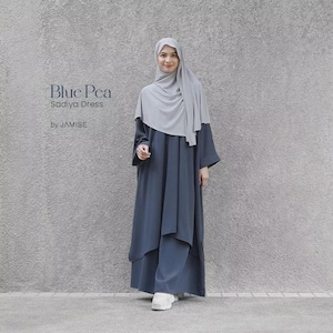 Puede incluir: Un vestido Sadiya azul oscuro de JAMISE con una silueta superpuesta y holgada, mangas largas y cuello alto, combinado con un hiyab gris claro. El texto Blue Pea Sadiya Dress by JAMISE está impreso en el lado izquierdo de la imagen.