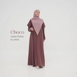 May include: A full-length brown dress and matching headscarf. The dress has long sleeves and a simple, flowing design. The text "Choco Laiqa Dress by JAMISE" is displayed on the left. The outfit is a modest and elegant style, suitable for various occasions.