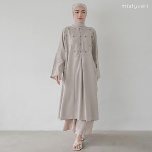 Può includere: Un abito beige a maniche lunghe con collo alto e chiusura a bottoni sul davanti. L'abito presenta dettagli floreali ricamati ed è abbinato a pantaloni beige coordinati e un copricapo. L'outfit è completato da sandali beige.