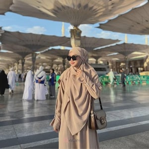 Op de afbeelding: Een vrouw draagt een beige hijab, zonnebril en een bijpassende jurk met lange mouwen. Ze draagt een schoudertas met patroon en een bruine leren riem. De achtergrond toont een grote buitenstructuur met open paraplu's en mensen.