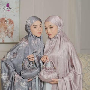 May include: Two women wearing matching headscarves and dresses with floral patterns. Each woman holds a small, matching handbag. The headscarves and dresses are in shades of grey and pink. The brand name "Jumana" is visible in the upper left and lower right corners.