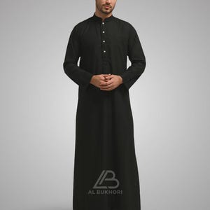 May include: A black long-sleeve thobe with a mandarin collar and button-down front. The thobe is floor-length and features the "AL BUKHORI" logo near the hem. The garment is made of a solid, dark fabric.
