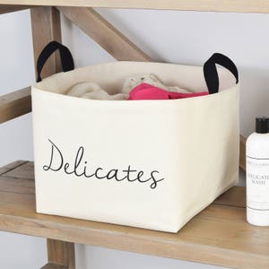 May include: A white canvas laundry basket with black handles and the word "Delicates" printed on the front. The basket is sitting on a wooden shelf.
