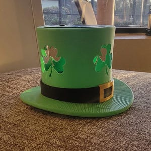 May include: A green St. Patrick's Day hat-shaped candle holder. The hat features shamrock cutouts, a black band, and a gold-coloured buckle. The brim is green and has a textured surface.