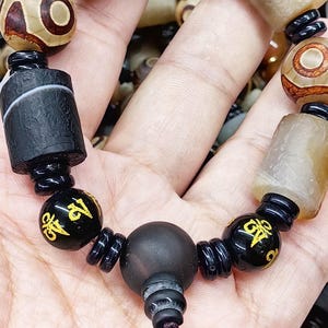 Peut inclure: Un bracelet composé de perles cylindriques et sphériques aux textures et couleurs variées. Il comprend des perles de style Dzi brunes et blanches à motifs, des perles cylindriques noir mat et des perles sphériques noires lisses avec des symboles dorés.