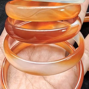 Pode incluir: Uma pilha de cinco pulseiras rígidas, lisas e polidas, feitas de pedra ágata translúcida. As pulseiras exibem um gradiente de cores que varia de um branco leitoso pálido no topo a tons profundos e vibrantes de laranja e castanho-avermelhado na base.