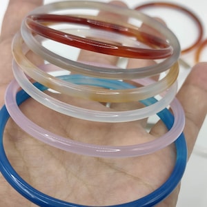 May include: A stack of thin, smooth, circular bangle bracelets in various translucent colors including deep red, soft grey, amber, milky white, pale pink, and vibrant blue, all featuring a polished finish and a rounded, uniform cross-section.
