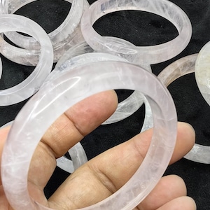 Puede incluir: Una colección de brazaletes de cuarzo rosa translúcido y pálido con un acabado suave y pulido. Cada brazalete presenta una forma circular con un perfil redondeado, mostrando la textura cristalina natural y las variaciones del color.