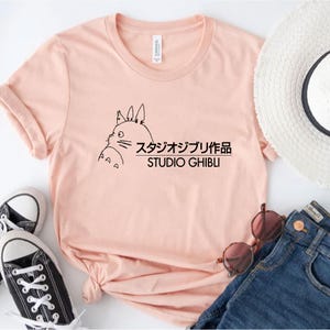 May include: A light pink t-shirt featuring a black outline illustration of a character from Studio Ghibli, with Japanese text and the English text "STUDIO GHIBLI". The shirt is styled with a pair of black sneakers, sunglasses, and blue jeans.