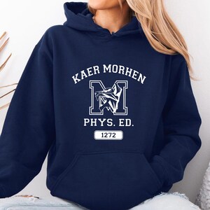 Puede incluir: Una sudadera con capucha azul marino que presenta un estampado gr&aacute;fico blanco en el pecho. El dise&ntilde;o incluye el texto KAER MORHEN, una letra M grande con un emblema de cabeza de lobo, el texto PHYS. ED. y un recuadro con el n&uacute;mero 1272.