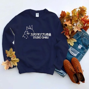 May include: Navy blue sweatshirt featuring a white outline of a cartoon character and the text "スタジオジブリ作品 STUDIO GHIBLI". The sweatshirt is paired with blue jeans and brown shoes.