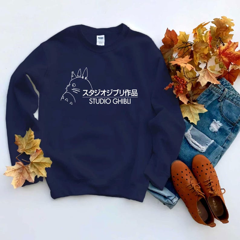 May include: Navy blue sweatshirt featuring a white outline of a cartoon character and the text "スタジオジブリ作品 STUDIO GHIBLI". The sweatshirt is paired with blue jeans and brown shoes.