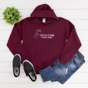 May include: Burgundy hoodie with a white graphic of a cartoon character and Japanese text. The hoodie has a front pocket and a drawstring hood. Black slip-on shoes and blue jeans are also in the image.