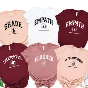 Puede incluir: Una colecci&oacute;n de seis camisetas de manga corta en varios colores, cada una con una palabra diferente en una fuente universitaria. Las palabras incluyen "SHADE", "EMPATH", "TELEPORTER", "FLASHER" y "HTECHNOPATH". Cada camiseta tambi&eacute;n tiene un peque&ntilde;o gr&aacute;fico y el texto "KEEPERS OF THE LOST CITIES".