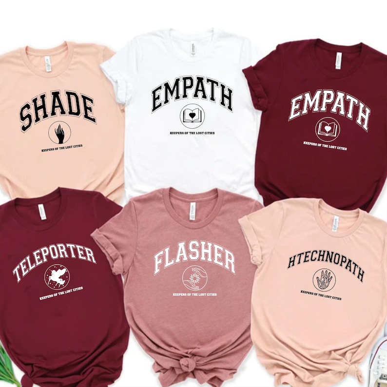 Puede incluir: Una colecci&oacute;n de seis camisetas de manga corta en varios colores, cada una con una palabra diferente en una fuente universitaria. Las palabras incluyen "SHADE", "EMPATH", "TELEPORTER", "FLASHER" y "HTECHNOPATH". Cada camiseta tambi&eacute;n tiene un peque&ntilde;o gr&aacute;fico y el texto "KEEPERS OF THE LOST CITIES".