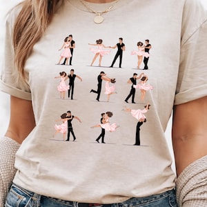 May include: Beige t-shirt featuring a series of illustrations of couples dancing. The dancers are depicted in black and white, with the women wearing pink dresses. The shirt has a round neckline and short sleeves.