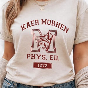 Puede incluir: Una camiseta de manga corta de color beige con texto en rojo oscuro que dice KAER MORHEN, PHYS. ED., y el n&uacute;mero 1272 dentro de un borde rectangular. Una letra M grande est&aacute; centrada con un gr&aacute;fico de cabeza de lobo estilizado.
