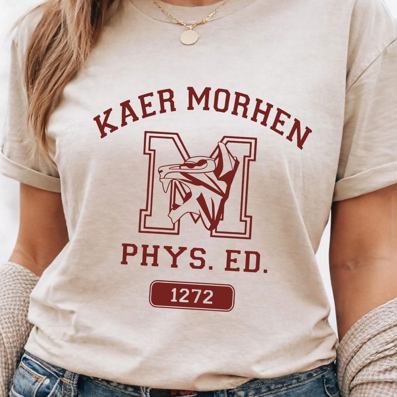 Puede incluir: Una camiseta de manga corta de color beige con texto en rojo oscuro que dice KAER MORHEN, PHYS. ED., y el n&uacute;mero 1272 dentro de un borde rectangular. Una letra M grande est&aacute; centrada con un gr&aacute;fico de cabeza de lobo estilizado.