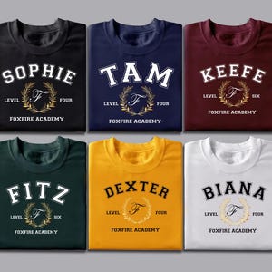 May include: Six folded t-shirts in various colors, each with a name, level, and "Foxfire Academy" text. The names include Sophie, Tam, Keefe, Fitz, Dexter, and Biana. Each shirt features a gold wreath design.