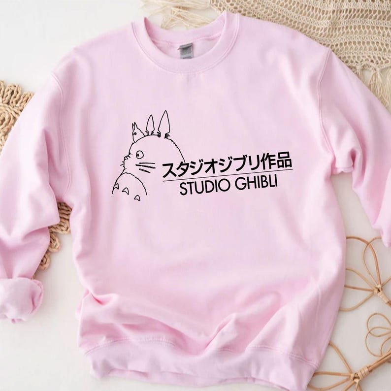 May include: A light pink sweatshirt featuring a black graphic of a cartoon character and Japanese text. The text reads "Studio Ghibli" in English and Japanese characters. The sweatshirt has long sleeves and a crew neck.