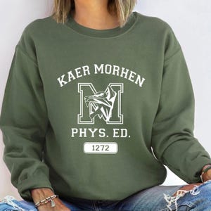 Puede incluir: Una sudadera de cuello redondo color verde oliva con texto serigrafiado en blanco que dice KAER MORHEN, una letra M grande con un gr&aacute;fico de cabeza de lobo, el texto PHYS. ED. y un recuadro rectangular con el n&uacute;mero 1272 centrado en el pecho.