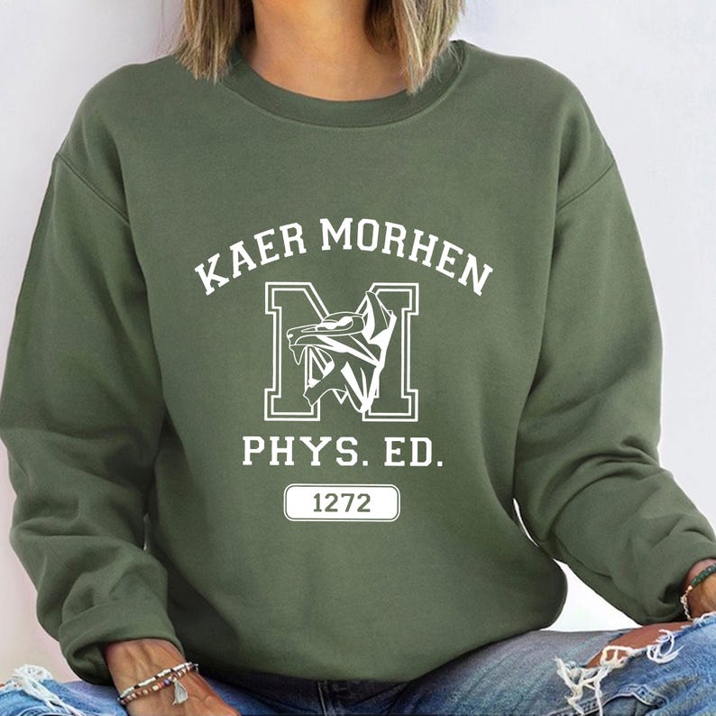 Puede incluir: Una sudadera de cuello redondo color verde oliva con texto serigrafiado en blanco que dice KAER MORHEN, una letra M grande con un gr&aacute;fico de cabeza de lobo, el texto PHYS. ED. y un recuadro rectangular con el n&uacute;mero 1272 centrado en el pecho.