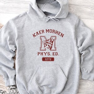 Puede incluir: Una sudadera con capucha gris claro que presenta un estampado gr&aacute;fico rojo oscuro en el pecho. El dise&ntilde;o incluye el texto KAER MORHEN, una letra M grande con un emblema de lobo, el texto PHYS. ED. y un recuadro con el n&uacute;mero 1272.