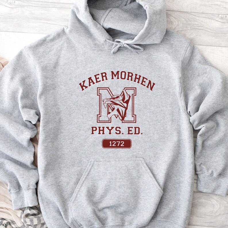 Puede incluir: Una sudadera con capucha gris claro que presenta un estampado gr&aacute;fico rojo oscuro en el pecho. El dise&ntilde;o incluye el texto KAER MORHEN, una letra M grande con un emblema de lobo, el texto PHYS. ED. y un recuadro con el n&uacute;mero 1272.