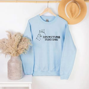 May include: A light blue sweatshirt featuring a black outline illustration of a cartoon character and Japanese text. The text reads "Studio Ghibli" in both Japanese characters and English. The sweatshirt is hanging on a wooden hanger.
