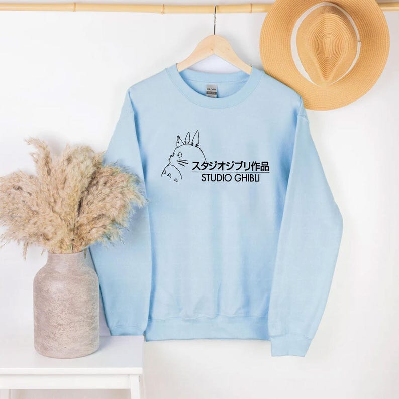 May include: A light blue sweatshirt featuring a black outline illustration of a cartoon character and Japanese text. The text reads "Studio Ghibli" in both Japanese characters and English. The sweatshirt is hanging on a wooden hanger.