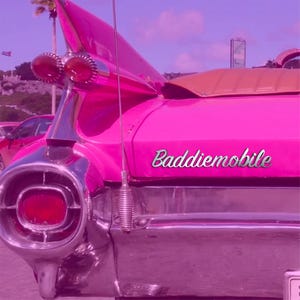 Könnte beinhalten: Nahaufnahme des Hecks eines klassischen rosa Autos mit Chromakzenten und einer markanten Heckflosse. Das Auto hat ein beiges Cabrio-Verdeck und das Wort "Baddiemobile" steht auf der Seite. Das Auto steht an einem sonnigen Tag draußen.