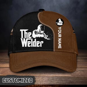 Puede incluir: Gorra de béisbol personalizada con una combinación de colores negro y marrón. La gorra presenta el texto "The Welder" y un gráfico de un soldador. Las palabras "YOUR NAME" están impresas verticalmente en el lateral. La palabra "CUSTOMIZED" está impresa en la visera.