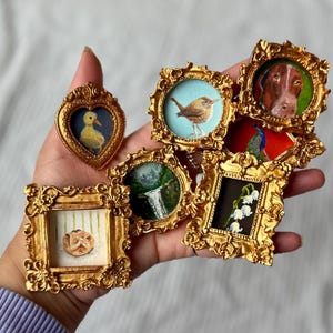 May include: A collection of miniature framed paintings with ornate gold frames. The paintings depict a duckling, a bird, a dog, a waterfall, a flower arrangement, and a peacock. The frames vary in shape, including heart and square.