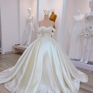 K&ouml;nnte beinhalten: Ein elfenbeinfarbenes Brautkleid aus Satin mit Off-Shoulder-Ausschnitt, weitem Rock und langer Schleppe. Das Oberteil ist mit zarter Stickerei versehen, und der Saum ist mit Spitze verziert. Eine goldene Krone sitzt auf dem Kopf der Schaufensterpuppe.