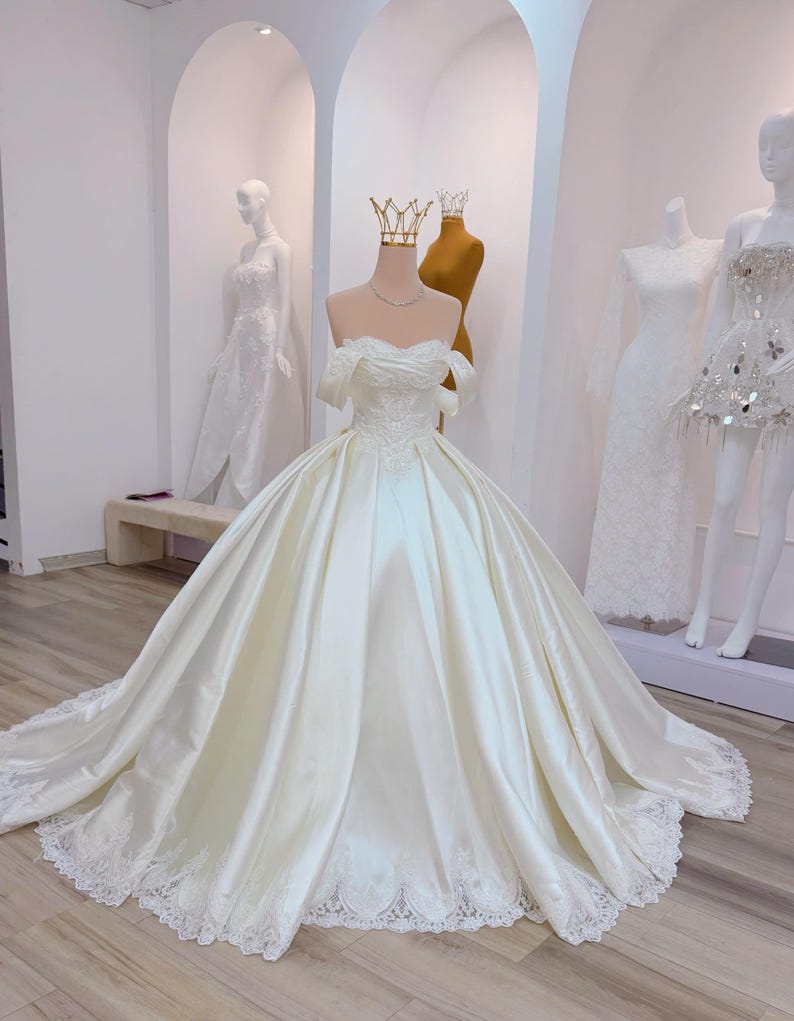 K&ouml;nnte beinhalten: Ein elfenbeinfarbenes Brautkleid aus Satin mit Off-Shoulder-Ausschnitt, weitem Rock und langer Schleppe. Das Oberteil ist mit zarter Stickerei versehen, und der Saum ist mit Spitze verziert. Eine goldene Krone sitzt auf dem Kopf der Schaufensterpuppe.