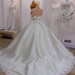 K&ouml;nnte beinhalten: Ein wei&szlig;es Brautkleid aus Satin mit langer Schleppe und Spitzenbesatz. Das Kleid hat ein schulterfreies Design und einen Korsettr&uuml;cken. Mannequins im Hintergrund zeigen weitere wei&szlig;e Kleider und eine goldene Krone.