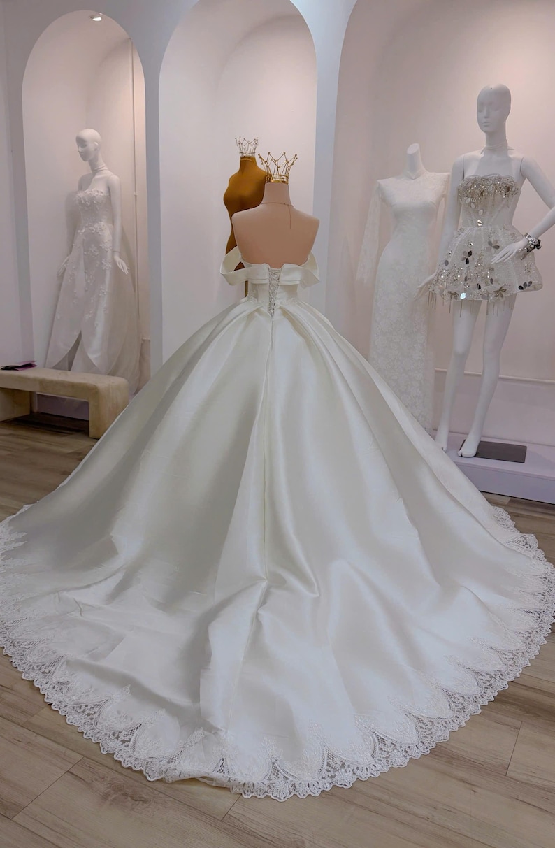 K&ouml;nnte beinhalten: Ein wei&szlig;es Brautkleid aus Satin mit langer Schleppe und Spitzenbesatz. Das Kleid hat ein schulterfreies Design und einen Korsettr&uuml;cken. Mannequins im Hintergrund zeigen weitere wei&szlig;e Kleider und eine goldene Krone.